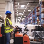 Woman warehouse worker operating forklift machine in large distribution warehouse center.